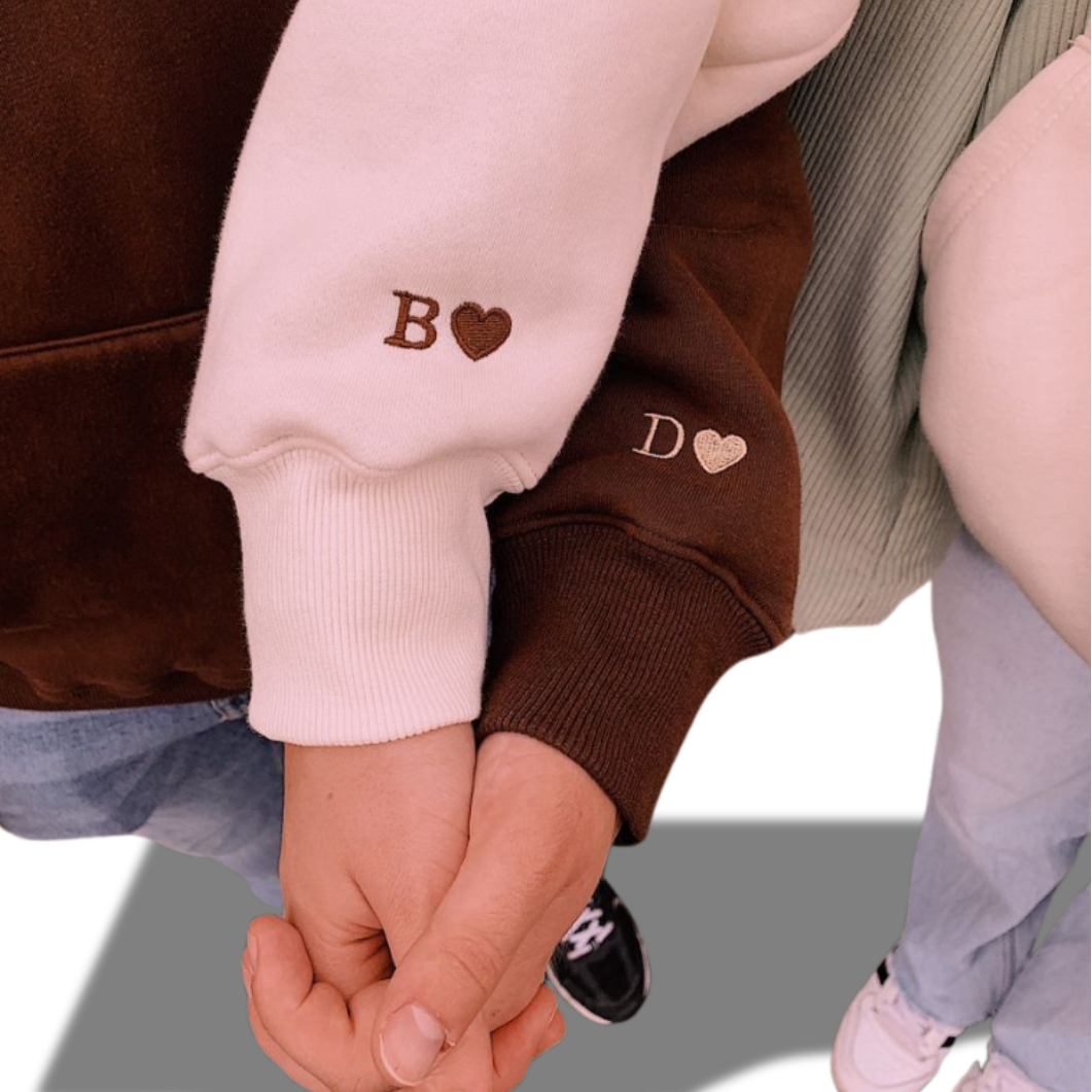 Two people holding hands wearing beige and brown hoodies with embroidered initials and hearts on the sleeves.